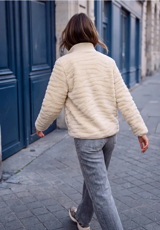 Femme en veste crème et pantalon gris marchant sur un trottoir pavé devant des portes bleues.