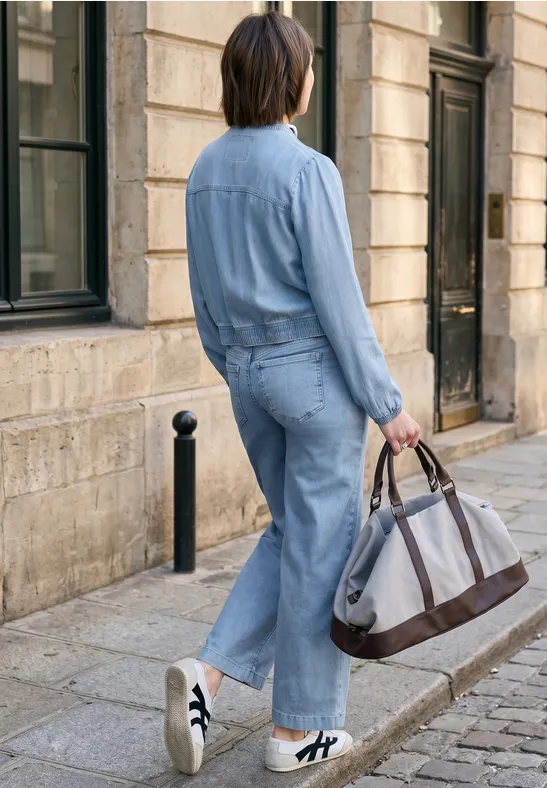 Persoon in lichtblauwe denim overall met grote tas loopt over een kasseienstraat langs een stenen gebouw.