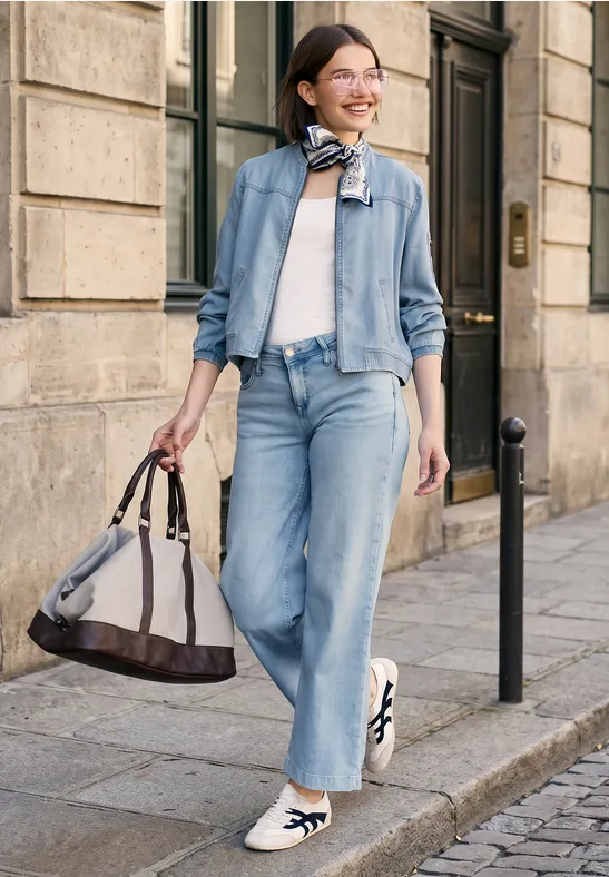 Vrouw in denim met reisttas en sjaal loopt voor een historische gevel