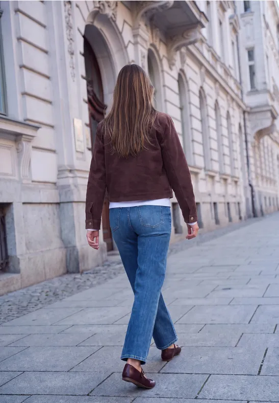 Frau in brauner Jacke und Jeans geht eine historische Straße entlang, gesehen von hinten.