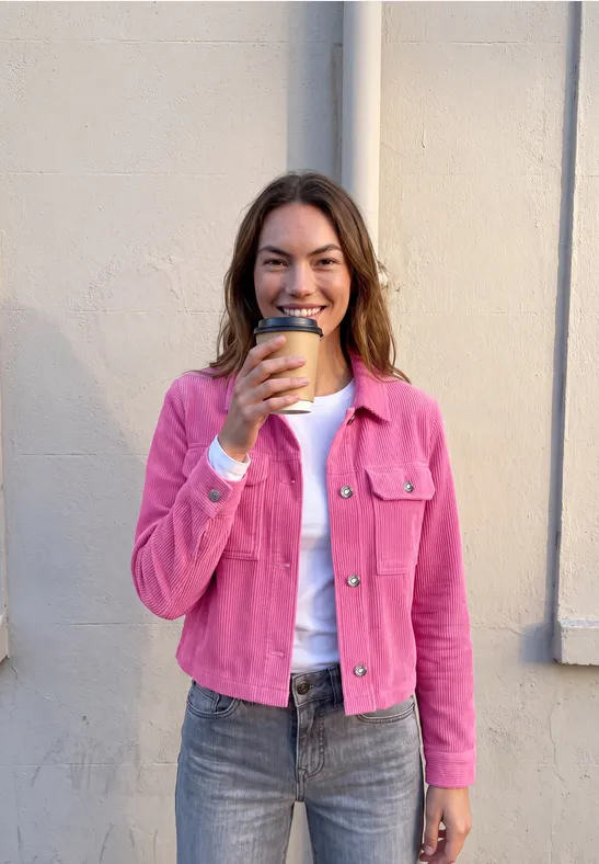 Eine lächelnde Frau in pinker Jacke und Jeans, die einen Kaffee in der Hand hält, vor einer hellen Wand.