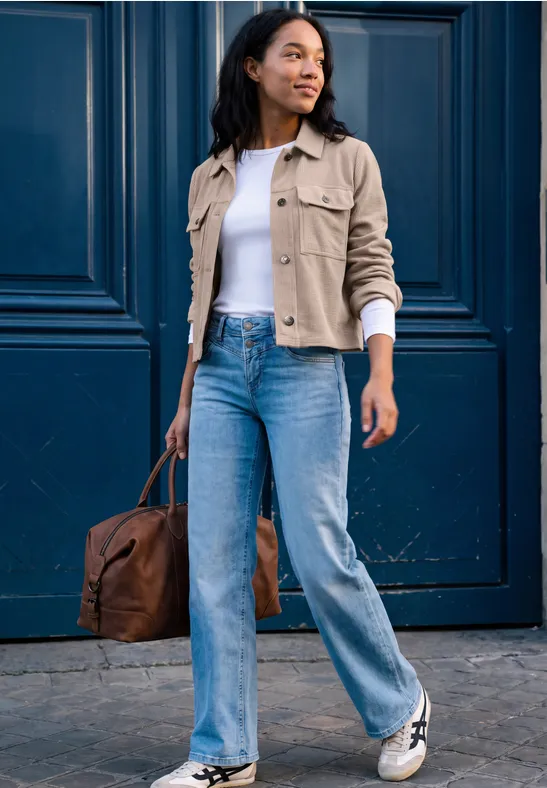 Eine junge Frau in Jeans, weißem Shirt und beiger Jacke trägt eine braune Tasche vor einer blauen Tür.