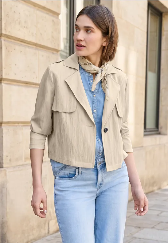 Vrouw in beige jas, spijkerbroek en sjaaltje voor een stenen muur.