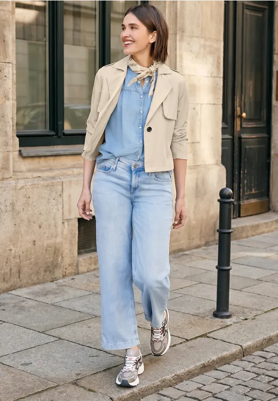 Vrouw in lichte spijkerbroek, blauw shirt, beige jasje en sneakers loopt op een geplaveid trottoir voor een gebouw.