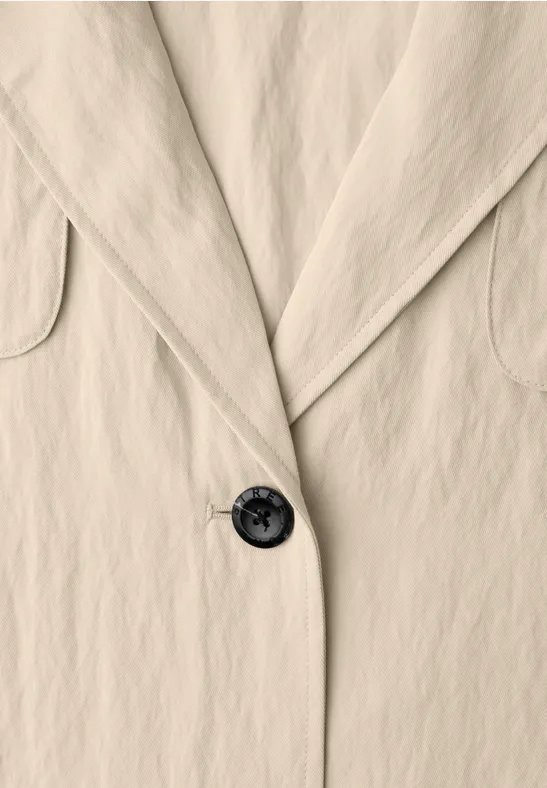 Close-up van een beige blazer met revers en een zwarte knoop.
