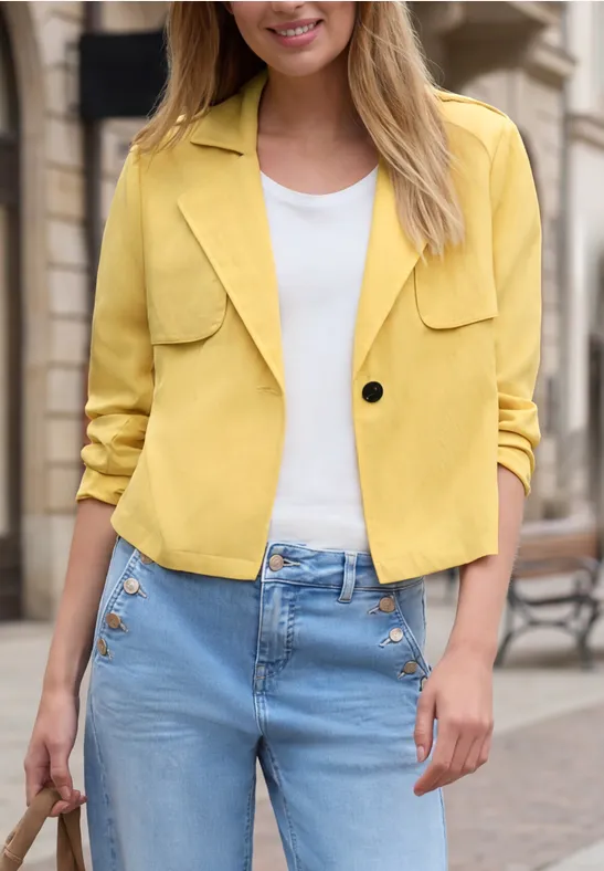 Frau mit gelber Jacke, weißem Shirt und Jeans mit goldenen Knöpfen hält eine Tasche.