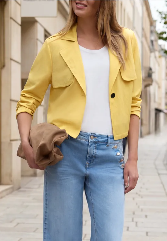 Frau in gelber Jacke, weißem Top und blauen Jeans, die einen Hut hält und auf einer gepflasterten Straße in der Stadt steht.