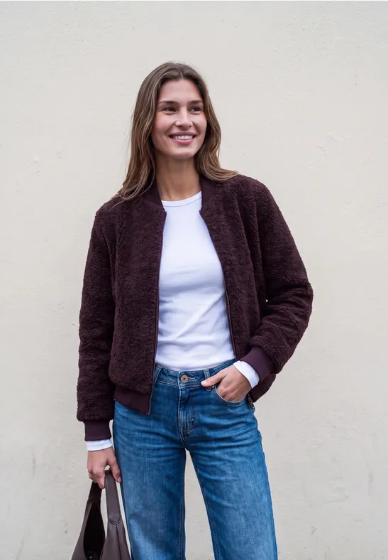 Eine lächelnde Frau in brauner Jacke, weißem Shirt und Jeans vor einer hellen Wand.