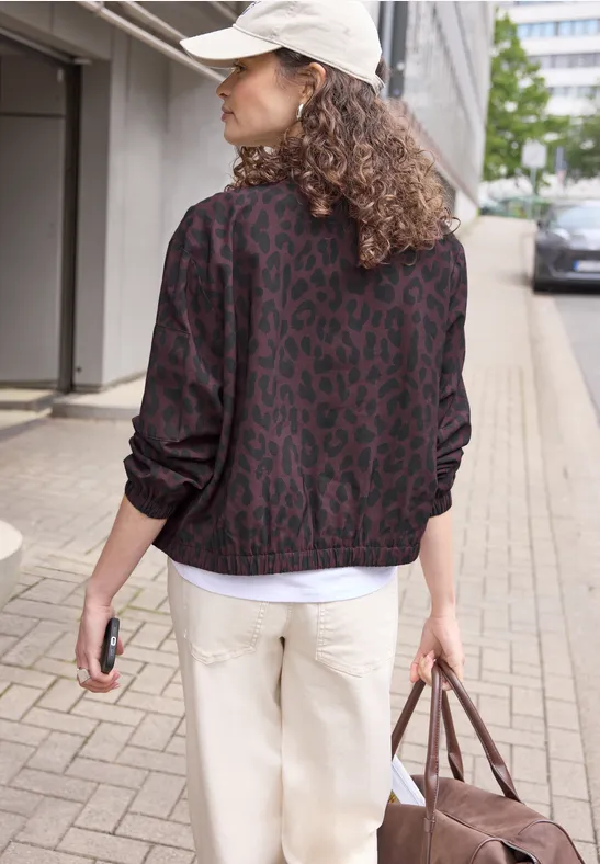 Rugaanzicht van een persoon met een patroonjas, witte pet en lichte broek, met een tas op het trottoir.