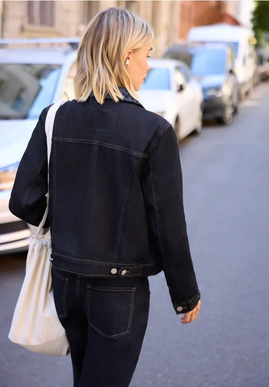 Eine blonde Frau trägt schwarze Denimjacke und -hose, läuft mit weißer Tasche eine städtische Straße entlang.