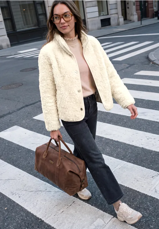 Frau trägt weiße Jacke, rosa Pullover, dunkle Jeans, weiße Sneaker, braune Reisetasche