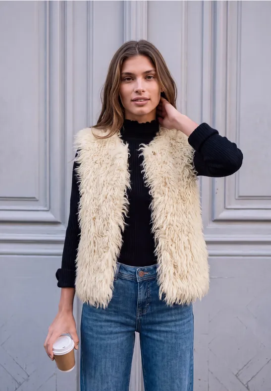Frau mit hellbraunen Haaren, schwarzem Langarmshirt, cremefarbener Teddyweste und blauen Jeans hält Kaffee.