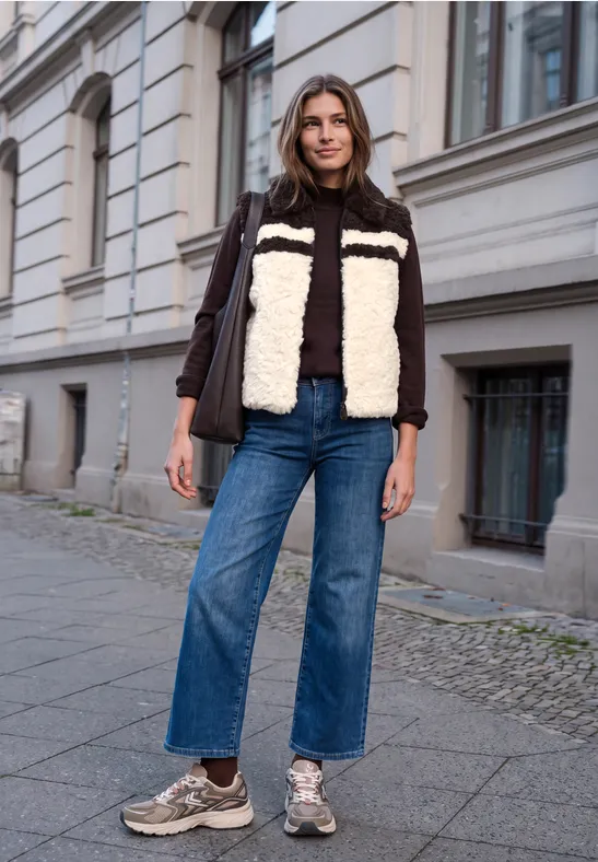 Frau steht auf Bürgersteig vor Gebäude, trägt zweifarbige Teddyjacke, weite Jeans und Sneakers.