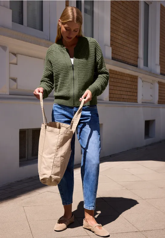 Frau in grüner Strickjacke, blauen Jeans und braunen Schuhen trägt eine Leinwandtasche und geht auf dem Gehweg.