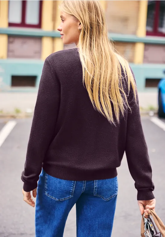 Blonde Frau von hinten in schwarzem Pullover und blauen Jeans, eine Tasche in der Hand, steht vor buntem Haus.