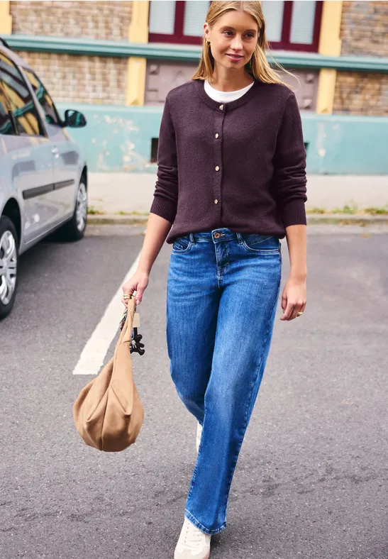 Eine Frau in Jeans und Strickjacke spaziert mit Tasche auf einem Parkplatz.