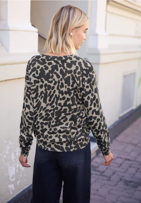 Frau von hinten in lockerem grünem Pullover mit schwarzem Leopardenmuster und dunklen Jeans auf dem Gehweg.