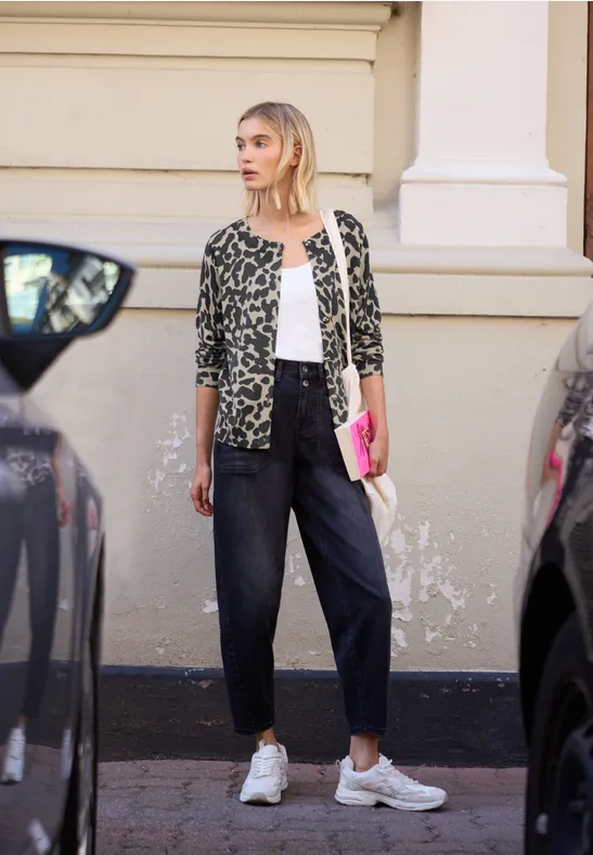 Junge Frau im Leoparden-Cardigan, weißem Top und Sneakers steht mit pinkem Heft zwischen parkenden Autos.