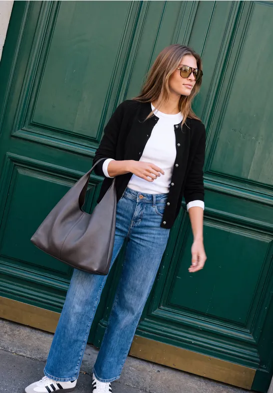 Stilvolle Frau in Jeans, weißem Shirt und Cardigan mit großer Ledertasche vor grünem Tor.