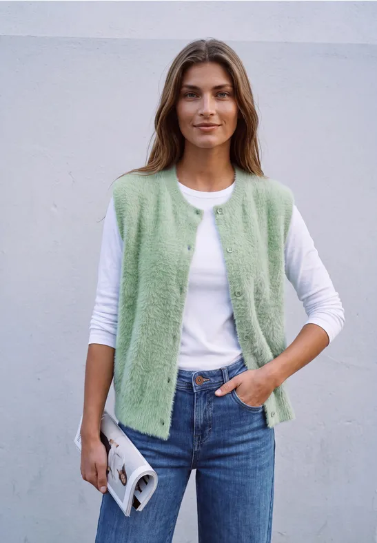 Junge Frau mit grüner Strickweste über weißem Shirt und Jeans, hält Magazin, grauer Hintergrund.