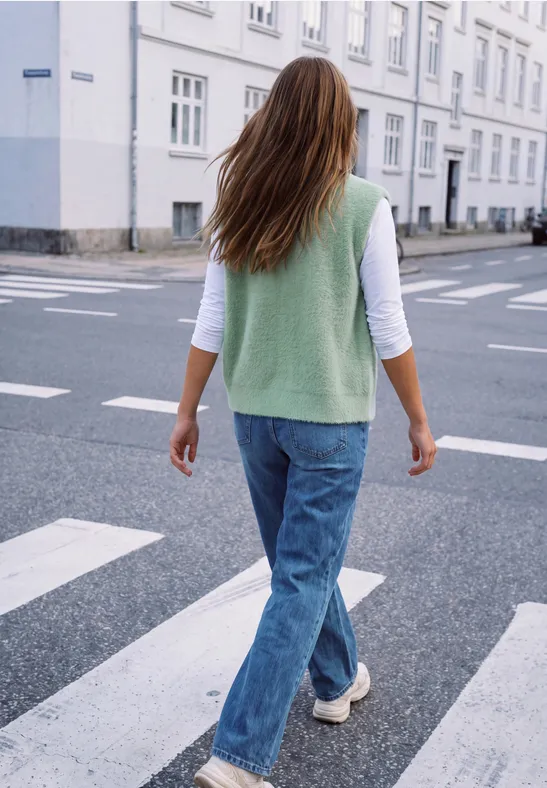 Eine Frau mit grünem Pullunder, weißen Shirt und Jeans überquert bei Tageslicht einen Zebrastreifen in der Stadt.