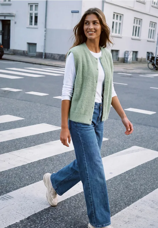 Frau in Jeans, weißem Shirt und grünem Strickwestchen überquert lächelnd bei sonnigem Wetter einen Zebrastreifen.