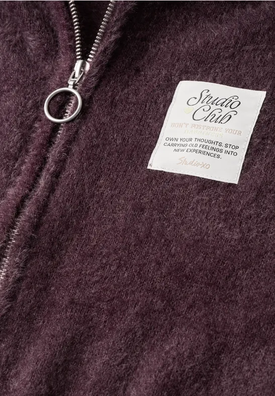 Nahaufnahme eines weinroten Fleece-Teils mit Reißverschlussring und weißem 'Studio Club'-Label.