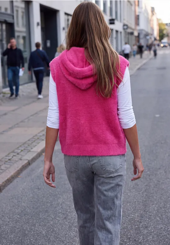Eine Frau in pinker Kapuzenweste und grauen Hosen geht eine belebte Straße entlang.