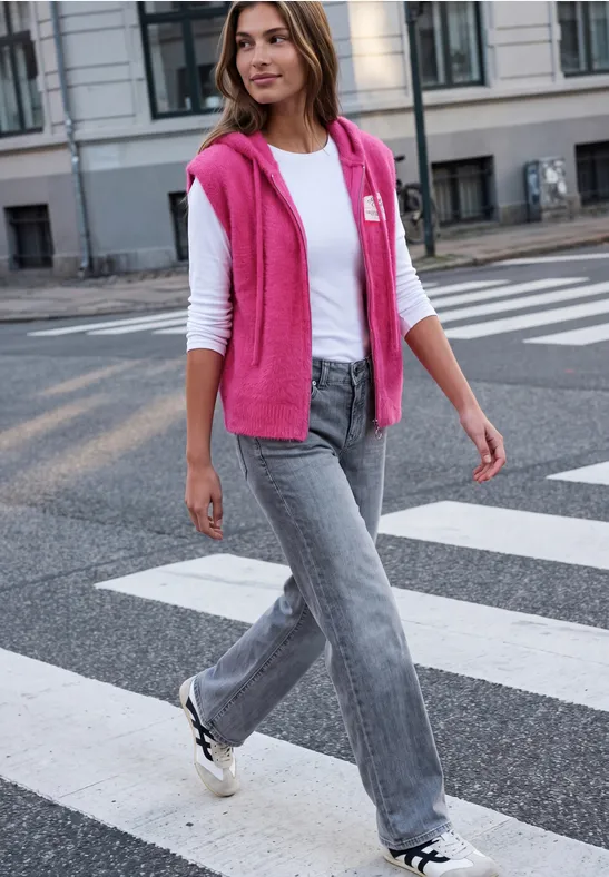 Frau in weißem Shirt, grauen Jeans und pinkem Pulli über den Schultern läuft über Zebrastreifen.