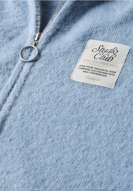 Hellblauer Fleece-Zippullover mit Ring-Reißverschluss und weißem Label mit Aufschrift 'Studio Club'.