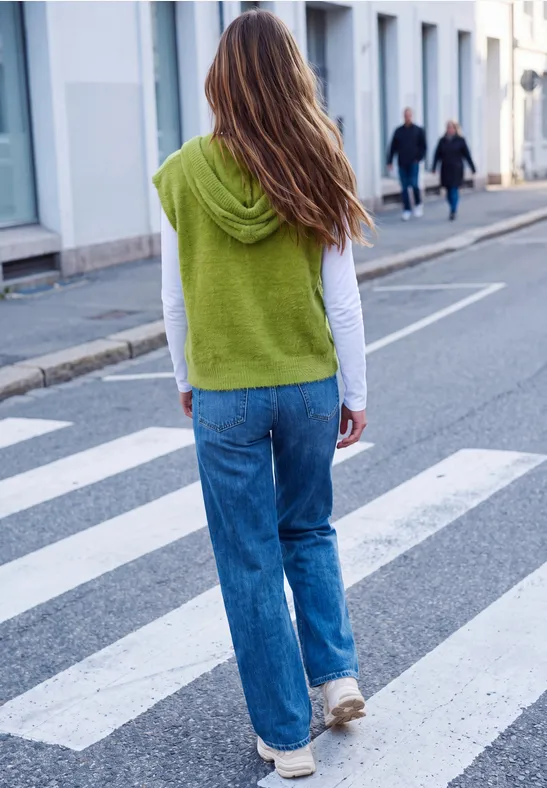 Vrouw in jeans en wit topje, groene trui over de schouders, loopt over een zebrapad.