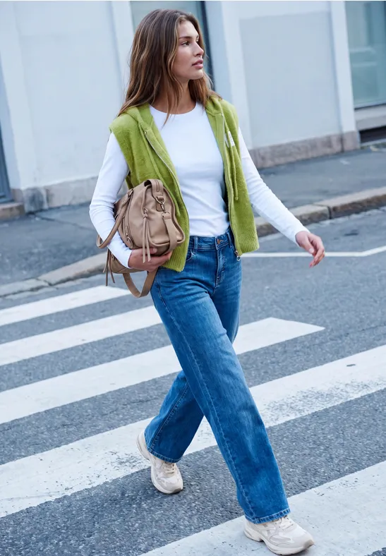 Een vrouw met wit shirt, groen vest, wijdere jeans en beige tas steekt een zebrapad over in de stad.