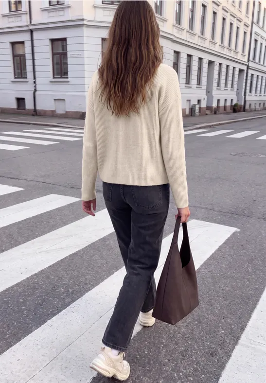 Eine Frau läuft auf einem Zebrastreifen in der Stadt, trägt beigen Pullover, schwarze Hose und weiße Sneaker.