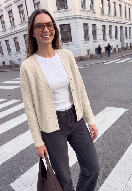 Junge Frau in Cardigan, weißem Shirt und Jeans mit Tasche an einem Zebrastreifen in der Stadt.