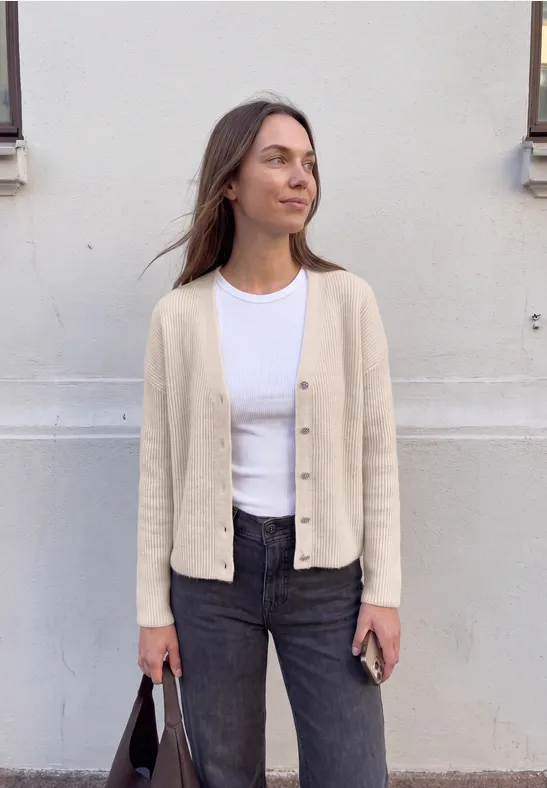 Junge Frau lehnt an weißer Wand, trägt beigen Cardigan, weißes Shirt und schwarze Jeans, hält schwarze Tasche.