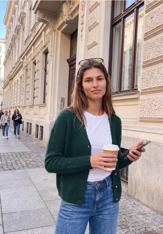 Junge Frau mit Kaffeebecher und Handy steht vor Gebäudefassade in urbaner Kopfsteinpflasterstraße.