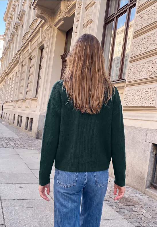 Eine Frau mit langen braunen Haaren in grünem Pullover und Jeans geht an historischer Fassade entlang.