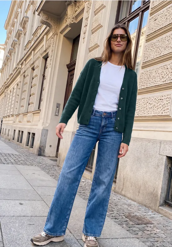Frau in grüner Strickjacke, weißem Shirt und weiter Jeans, lässig vor historischer Hausfassade.