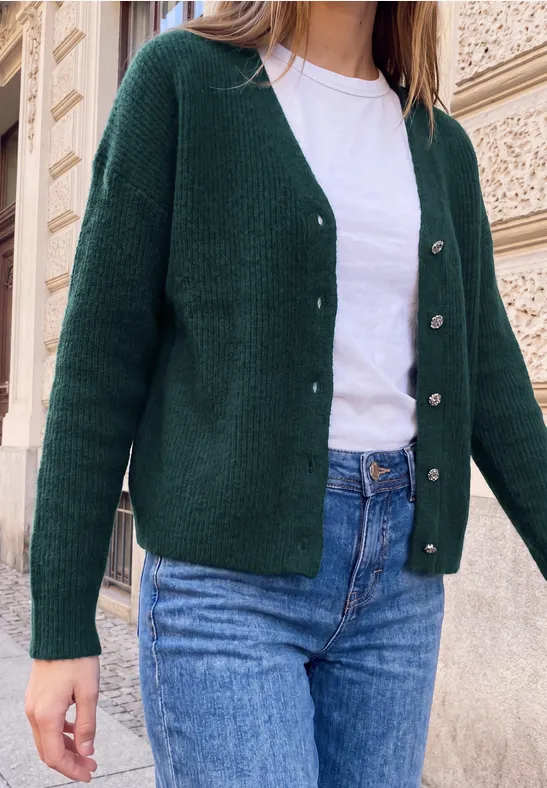 Frau trägt grünen Cardigan, weißes Shirt und blaue Jeans vor einer steinernen Hausfassade.