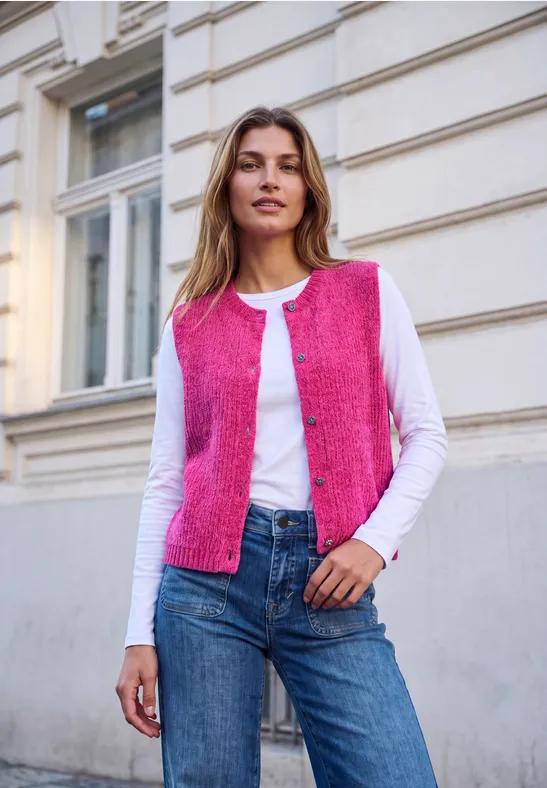 Frau in weißem Langarmshirt, pinker Weste und Jeans steht vor einer Hausfassade.