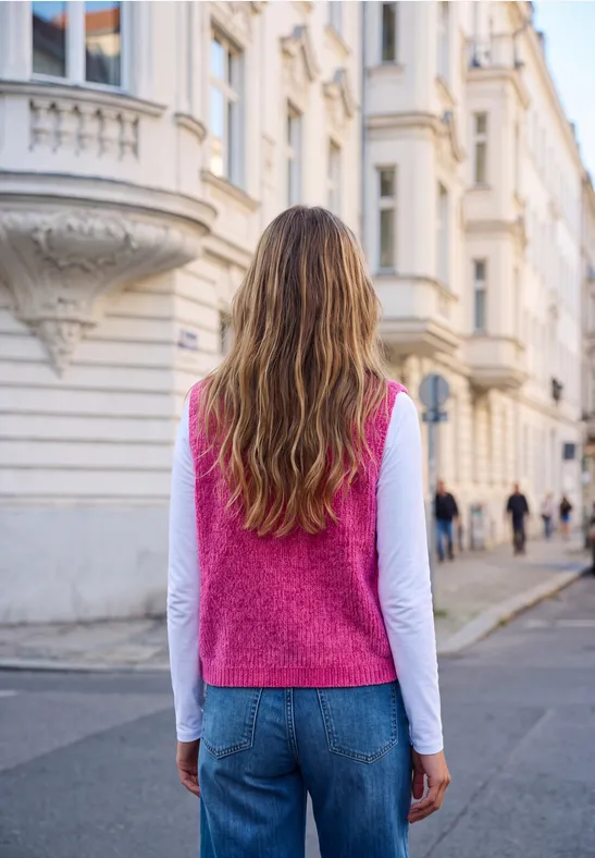 Eine Frau mit rosa Pullunder und blauer Jeans steht von hinten auf einer ruhigen Stadtstraße vor historischen Gebäuden.