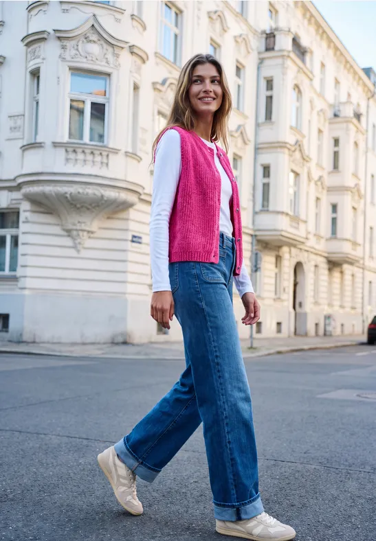 Eine Frau in pinkem Cardigan und Jeans läuft lächelnd durch eine elegante Altstadtstraße.