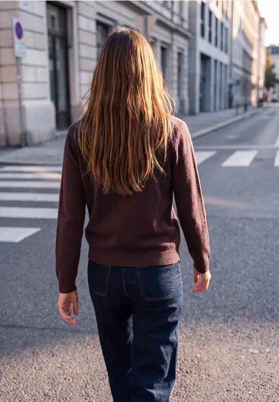 Eine Person mit langen Haaren in Jeans und Pullover geht über einen Zebrastreifen in einer ruhigen Straße.