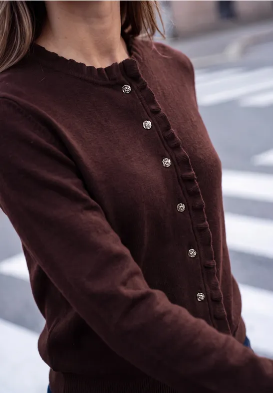 Eine Person trägt ein braunes Strickjäckchen mit Knöpfen vor städtischem Hintergrund.