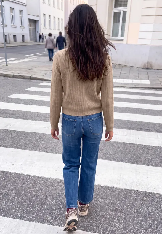 Eine Frau in braunem Pullover, blauen Jeans und Sneakers überquert einen Zebrastreifen in einer Stadt.