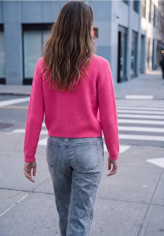 Frau mit langen braunen Haaren geht in pinkem Pullover und grauen Jeans über Zebrastreifen in der Stadt.