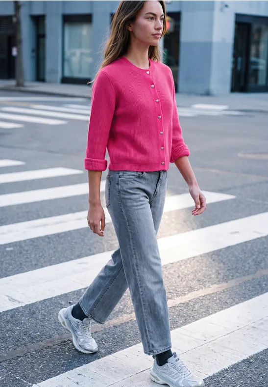 Eine Frau in pinkem Cardigan, grauen Jeans und weißen Sneakern überquert einen Zebrastreifen in der Stadt.