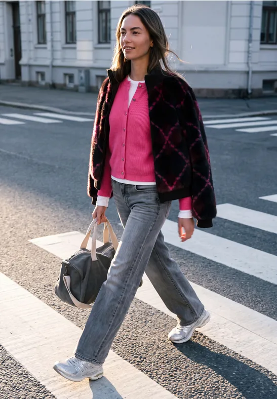 Junge Frau überquert selbstbewusst einen Zebrastreifen, trägt pinken Pullover, graue Hose und hält eine Reisetasche.