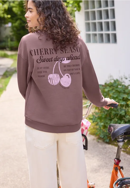 Femme aux cheveux bouclés en sweat à capuche marron avec motif cerises, debout à côté d’un vélo rouge en plein air.