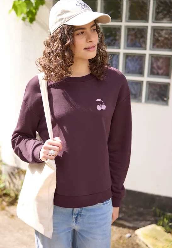 Person mit lockigem Haar und Basecap steht vor einem Haus, trägt lila Sweatshirt mit Kirschmotiv und Jeans